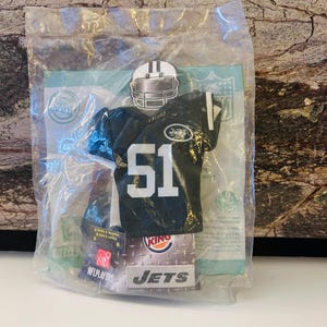 Puede incluir: Un uniforme de fútbol americano en miniatura de los Jets de Nueva York y un casco, empaquetados en una bolsa de plástico transparente. La camiseta negra lleva el número 51 en blanco, con el logotipo de los Jets. El embalaje incluye el logotipo de Burger King y el texto "NFL PLAYERS".