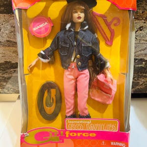 Może przedstawiać: Zapakowana lalka Bianca z serii Girl Force firmy Jakks Pacific. Lalka ma na sobie dżinsową kurtkę z wykończeniem ze sztucznego futra, różowe spodnie i różową torebkę. Opakowanie zawiera napis "SO WILD" i ostrzeżenie o małych częściach.