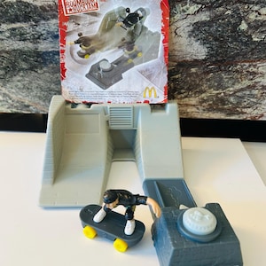 Op de afbeelding: Een grijs plastic Tony Hawk Boom Boom Huck Jam speelset met een miniatuur skateboarder figuur. De set bevat een helling, een platform en een klein skateboard. De verpakking is zichtbaar op de achtergrond.