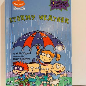 Puede incluir: Un libro infantil titulado "Stormy Weather" del Nickelodeon Book Club. La portada presenta a los personajes de Rugrats bajo un paraguas bajo la lluvia. El libro tiene un fondo azul con el título en letras grandes y verdes.