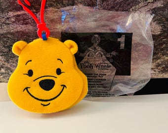 Könnte beinhalten: Ein gelber Winnie-the-Pooh-Plüschkopf mit einem schwarzen, lächelnden Mund und Augen, befestigt an einem roten Clip. Hinter dem Plüschtier befindet sich eine Plastiktüte mit Text.