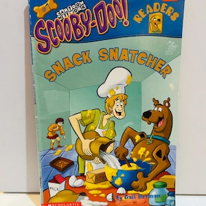 Puede incluir: Libro infantil titulado "Scooby-Doo! Snack Snatcher". La portada presenta a los personajes de dibujos animados Scooby-Doo y Shaggy cocinando. El fondo es azul con las palabras "Cartoon Network" y "Readers".
