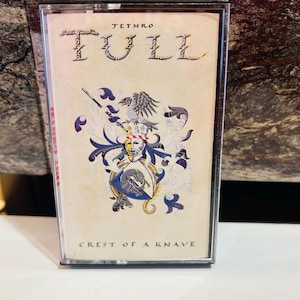 Puede incluir: Una cinta de casete de Jethro Tull "Crest of a Knave". La caja transparente de la cinta muestra la portada con el nombre de la banda y el título del álbum en una fuente estilizada, y un diseño de escudo en azul, dorado y blanco.