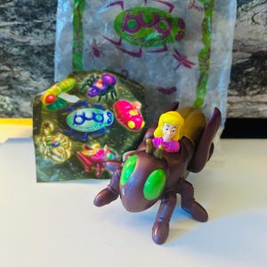 May include: A brown and gold toy insect with green eyes and a blonde figure riding on its back. A colorful card with various toy insects is behind the toy insect. A clear plastic bag with the word "Bug" is in the background.