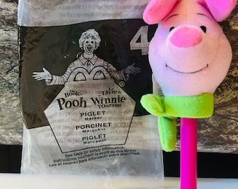 Könnte beinhalten: Ein Piglet-Marker-Spielzeug mit einem rosa Plüschkopf, rosa Ohren und einem grünen Kragen. Der Marker hat einen leuchtend pinkfarbenen Stiel. Die Verpackung ist hinter dem Spielzeug sichtbar, mit dem Text "Pooh Winnie" und "PIGLET Marker".