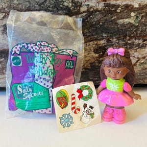 Könnte beinhalten: Eine Plastikpuppe mit braunen Haaren und einem rosa Kleid. Die Puppe hält einen grün-weißen Pullover mit einem Schneeflockenmuster. Die Puppe ist Teil einer McDonald's Happy Meal "Totally Toy Holiday" Aktion mit Sally Secrets. Die Puppe kommt mit einem Bogen Aufkleber mit einem Schneemann, einem Zuckerstangen, einer Schneeflocke und einem Kranz.