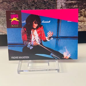 May include: A ProSet Super Stars Musicards trading card featuring Yngwie Malmsteen. The card has a pink and blue background with a photo of the musician playing guitar. The card is displayed on a clear plastic stand.