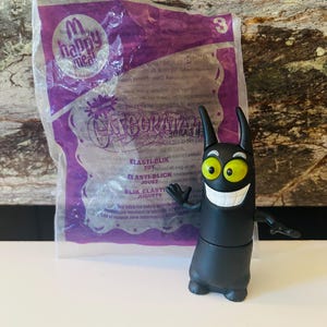 May include: A black Catscratch toy figure with yellow eyes and a wide white grin, standing in front of a purple Happy Meal bag. The bag has the words "Happy Meal" and "Catscratch" printed on it.