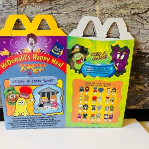 May include: Two McDonald's Happy Meal boxes. One is yellow with a white handle, featuring Ronald McDonald and the text "Monster Crazy Bones". The other is green with a white handle, with a witch and a purple monster. The boxes have colorful graphics.