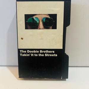 May include: An 8-track tape of The Doobie Brothers' album, "Takin' It to the Streets." The album cover features a close-up of a person wearing sunglasses. The text "The Doobie Brothers Takin' It to the Streets" is printed in white on the black tape.