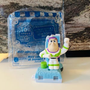 Puede incluir: Figura de juguete de Buzz Lightyear de Toy Story 4, en una bolsa de pl&aacute;stico transparente. La figura es blanca, verde y morada, con una base azul. La bolsa tiene el logotipo de Toy Story 4 y el texto "Scan Ticket Inside".