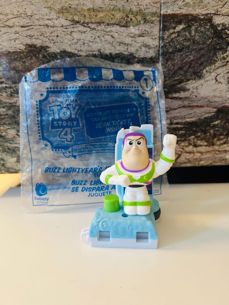 Puede incluir: Figura de juguete de Buzz Lightyear de Toy Story 4, en una bolsa de pl&aacute;stico transparente. La figura es blanca, verde y morada, con una base azul. La bolsa tiene el logotipo de Toy Story 4 y el texto "Scan Ticket Inside".