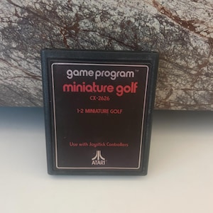 Peut inclure: Cartouche de jeu Atari noire avec le texte "game program" et "miniature golf" en blanc. La cartouche comporte également le texte "CX-2626" et "1-2 MINIATURE GOLF" en blanc. Le texte "Use with Joystick Controllers" et le logo Atari sont en blanc.