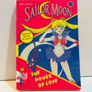 May include: A vintage Sailor Moon comic book with a vibrant pink cover. The cover features the title "SAILOR MOON" in large, stylized letters, along with an illustration of a Sailor Moon character and a black cat. The text "THE POWER OF LOVE" is also visible.