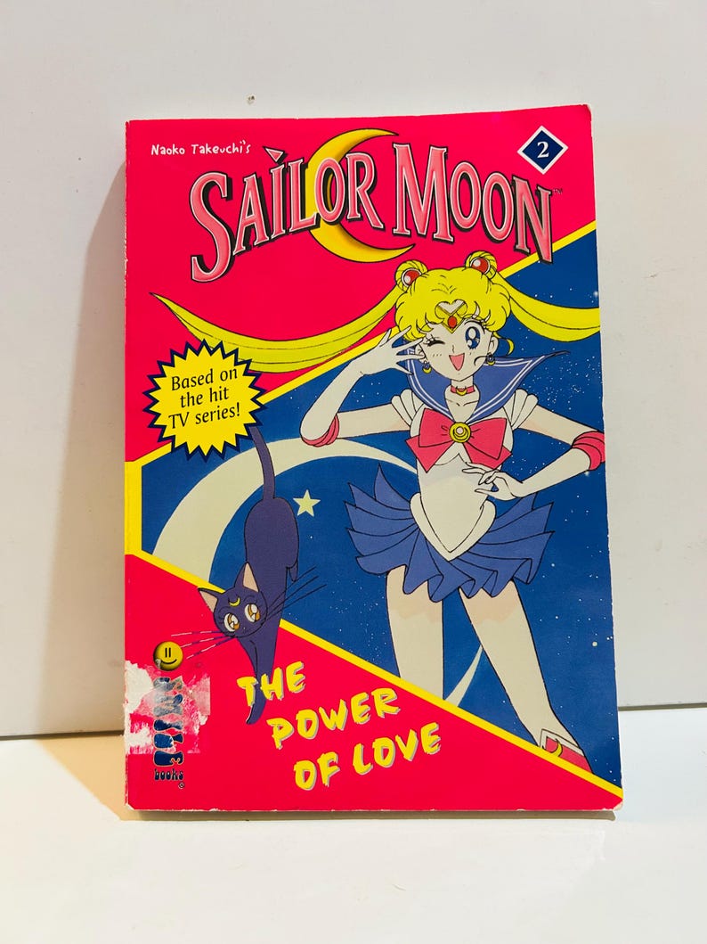 May include: A vintage Sailor Moon comic book with a vibrant pink cover. The cover features the title "SAILOR MOON" in large, stylized letters, along with an illustration of a Sailor Moon character and a black cat. The text "THE POWER OF LOVE" is also visible.