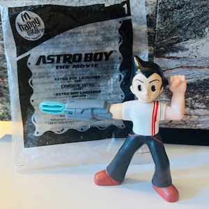 May include: Astro Boy action figure from the movie, with a white shirt, red accents, black pants, and red shoes. The figure is holding a gray and blue launcher. The packaging reads "Astro Boy The Movie".