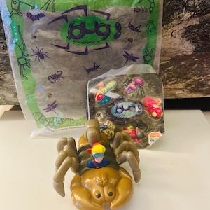 May include: A brown plastic toy scorpion with a small figure riding on its back. The toy is part of a "Bug Riders" set, with a clear plastic bag containing additional bug-themed toys in the background. The packaging features the "Bug Riders" logo and insect illustrations.