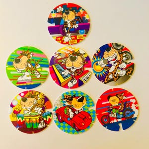 May include: A collection of seven colorful, circular game pieces featuring a cartoon cheetah character. Each piece depicts the cheetah in various poses, such as playing musical instruments, driving a car, and riding a motorcycle. The pieces have a unique shape with cut-out sections.