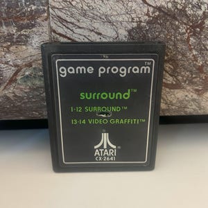Puede incluir: Cartucho de juego Atari negro con texto verde que dice "game program", "surround", "1-12 SURROUND", "13-14 VIDEO GRAFFITI" y "ATARI CX-2641".