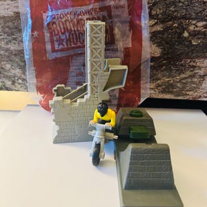 Può includere: Un set di giocattoli in plastica grigia con una moto e un pilota in miniatura, una rampa e una facciata di un edificio. La confezione recita "Tony Hawk's Boom Boom Huck". Il pilota indossa una maglietta gialla e un casco nero.