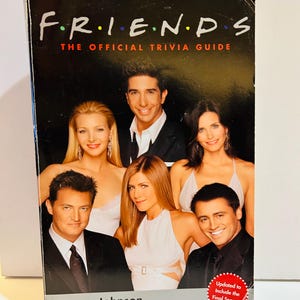 Puede incluir: Un libro titulado "FRIENDS THE OFFICIAL TRIVIA GUIDE" con el elenco de la serie de televisión. La portada tiene un fondo negro con el logotipo del programa en letras coloridas. El libro es de Lauren Johnson e incluye un prólogo de James Michael Tyler.