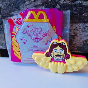 Könnte beinhalten: Ein Vintage Birdie-Spielzeug-Sound-Maker von McDonald's. Das gelb-rosa Kunststoffspielzeug zeigt eine Cartoon-Ente mit rosa Kleid und roten Haaren. Die Verpackung ist rosa und gelb mit dem McDonald's-Logo und der Birdie-Figur.