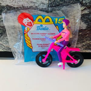 May include: A pink Barbie figurine on a pink bicycle, packaged in a clear plastic bag. The packaging features the McDonald's logo and text that reads "BICYCLING BARBIE FIGURINE". The Barbie figurine is wearing a pink and purple outfit.