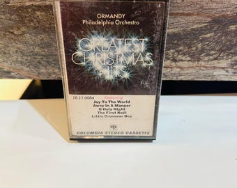 May include: A vintage cassette tape featuring "Greatest Christmas Hits" by Ormandy and the Philadelphia Orchestra. The cassette has a dark background with the album title in a bright, stylized font. The tracklist includes "Joy To The World" and "Little Drummer Boy."