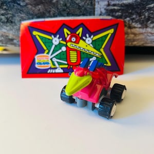 May include: A pink and green toy car with black wheels and white rims. A red sticker with a cartoon dinosaur and the text "Burger King" is in the background. The toy car has a green beak and blue accents.