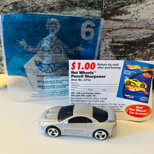 May include: A silver Hot Wheels toy car, a Mitsubishi Eclipse model, with black windows and wheels. A blue package with the number 6 and a Hot Wheels pencil sharpener are also visible.