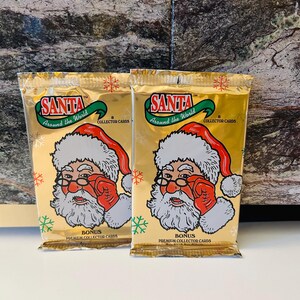 May include: Two gold-colored Santa Around the World collector card packs. Each pack features Santa Claus in a red hat and gloves, with the text "SANTA" and "Around the World". The packs are labeled "BONUS PREMIUM COLLECTOR CARDS".
