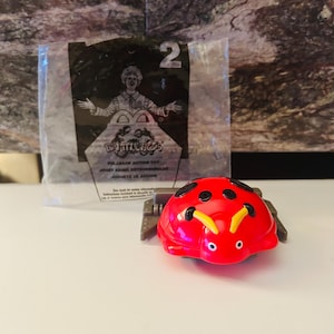May include: A red ladybird toy with black spots and yellow antennae, on a grey base. Behind it is a clear plastic bag with the number 2 and a clown graphic.