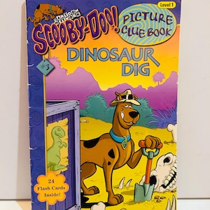 Puede incluir: Un libro de pistas ilustradas de Scooby-Doo! "Dinosaur Dig". La portada presenta a Scooby-Doo con una pala, cavando cerca de un esqueleto de dinosaurio. El libro incluye 24 tarjetas didácticas y es de nivel 1.