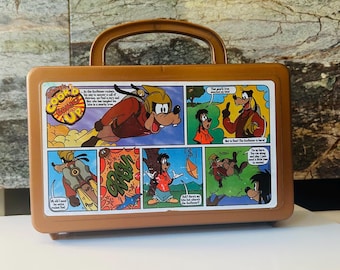May include: Brown vintage lunchbox featuring a comic strip with Goofy characters. The lunchbox has a brown handle and a rectangular shape. The comic strip has colorful panels with speech bubbles and action scenes.