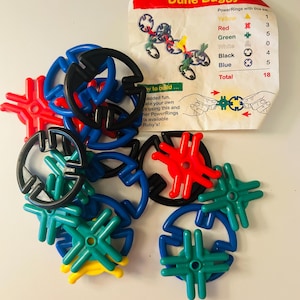 Puede incluir: Un conjunto de juguetes de construcción PowerRings Dune Buggy de colores. El juego incluye piezas amarillas, rojas, verdes, negras y azules, junto con una tarjeta impresa que muestra la lista de piezas y un diseño de buggy de muestra. El total es de 18 piezas.