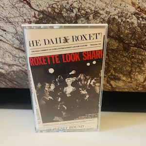 Puede incluir: Una cinta de casete con una foto en blanco y negro de una banda en la parte delantera. El texto en la cinta de casete dice "HE DAILY ROXET", "THE DAILY COMMENT ON GLOBAL ENTERTAINMENT AND POP CULTURE", "ROXETTE LOOK SHARE", "HEAVENLY BOUND..." y "Wednesday, Oct".