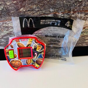 May include: A red and yellow ESPN 4 football electronic game featuring Michael Vick. The game has a digital display and is surrounded by footballs. The packaging includes the McDonald's logo.