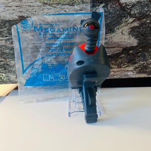 May include: A grey and red robot toy from a Happy Meal, with a round head and a red eye, and a flexible neck. The toy is on a clear plastic stand, with a partially visible Happy Meal bag in the background.