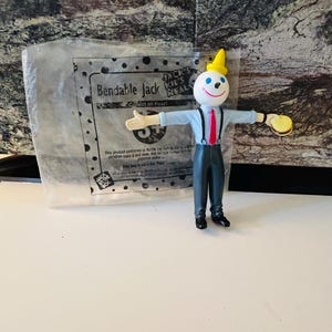 May include: A Bendable Jack in the Box kids meal toy figurine. The figure has a white head with a smiling face, a yellow hat, and a red tie. It is wearing a light blue shirt, gray pants, and black shoes. The figure is holding a small burger. A plastic bag is behind the figure.