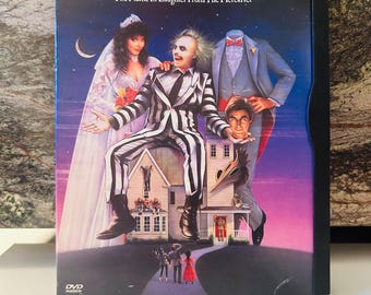 Pode incluir: Capa de DVD do filme Beetlejuice. A capa apresenta Michael Keaton em um terno listrado em preto e branco, com outros personagens e uma casa. O título está em letras douradas em um fundo azul escuro.