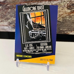 May include: A vintage concert poster for the Fillmore West in San Francisco, California. The poster features a black and white illustration of a man looking out a window. The poster lists the performers: R.R. King, Aum, Frost, Santana, Taj Mahal, and Eurovies. The dates of the concert are July 11-13, 1969.