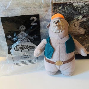 Könnte beinhalten: Ein Plüschtier von Big Al aus den Country Bears, mit einer tealfarbenen Weste, einem braunen Gürtel und einer orangefarbenen Kappe. Das Spielzeug stammt aus einem McDonald's Happy Meal, mit der Nummer "2" auf der Verpackung.