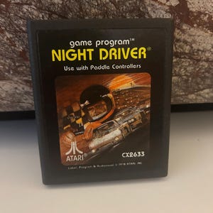 May include: A black Atari game cartridge with a yellow label. The label reads "game program™ NIGHT DRIVER® Use with Paddle Controllers". The cartridge has a picture of a race car driver and a sunset on the front. The text "ATARI® CX2633 Label, Program & Audiovisual © 1978 ATARI INC" is printed on the bottom of the label.