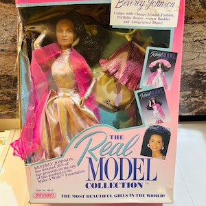 Op de afbeelding: Een pop uit de "The Real Model Collection" van Beverly Johnson in de doos. De pop draagt een gouden en roze jurk met een roze doorschijnende cape. De doos is roze en wit met de tekst "The Most Beautiful Girls in the World!"