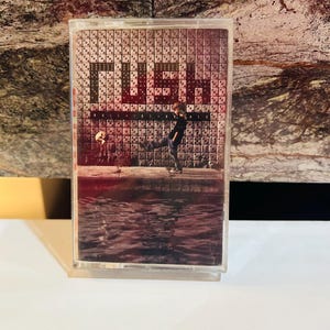 May include: A vintage cassette tape with the word "RUSH" in large letters. The cover art features a person in silhouette against a grid of dice. The background has a water reflection effect. The cassette is in a clear plastic case.