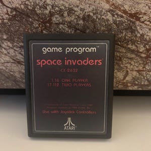 May include: A black Atari game cartridge with the text "game program" and "space invaders" in white. The cartridge also includes the text "CX-2632", "1-16 ONE PLAYER", "17-112 TWO PLAYERS", "Program contents © 1980 ATARI, INC." and "Use with Joystick Controllers". The Atari logo is in white on a black background.
