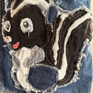 May include: A denim patch featuring a cartoon skunk design. The skunk is primarily black and white with blue eyes and a red mouth. The patch is sewn onto a blue denim background, with frayed edges for a distressed look.