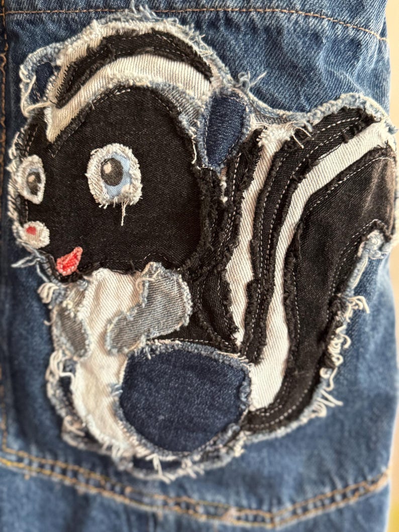 May include: A denim patch featuring a cartoon skunk design. The skunk is primarily black and white with blue eyes and a red mouth. The patch is sewn onto a blue denim background, with frayed edges for a distressed look.