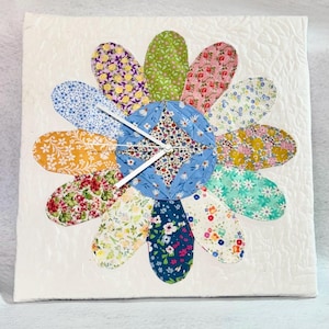 May include: A square, quilted wall clock with a flower design. The clock face is a patchwork of floral fabrics in various colors, including blue, yellow, pink, and green. White clock hands indicate the time. The background is white.