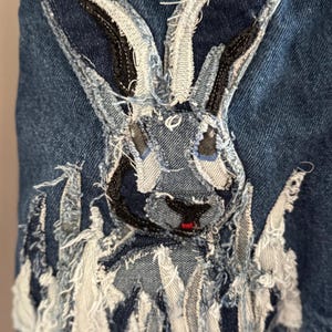 May include: Close-up of a denim jacket detail featuring a rabbit applique. The rabbit is constructed from various denim scraps in shades of blue, white, and gray, with frayed edges. The rabbit has black eyes and a red nose.